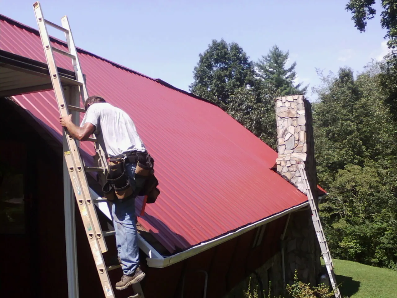 Roofer on a ladder beside a red metal roof and stone chimney — Soffit & Fascia Repair by Roofing Brothers in Newbern